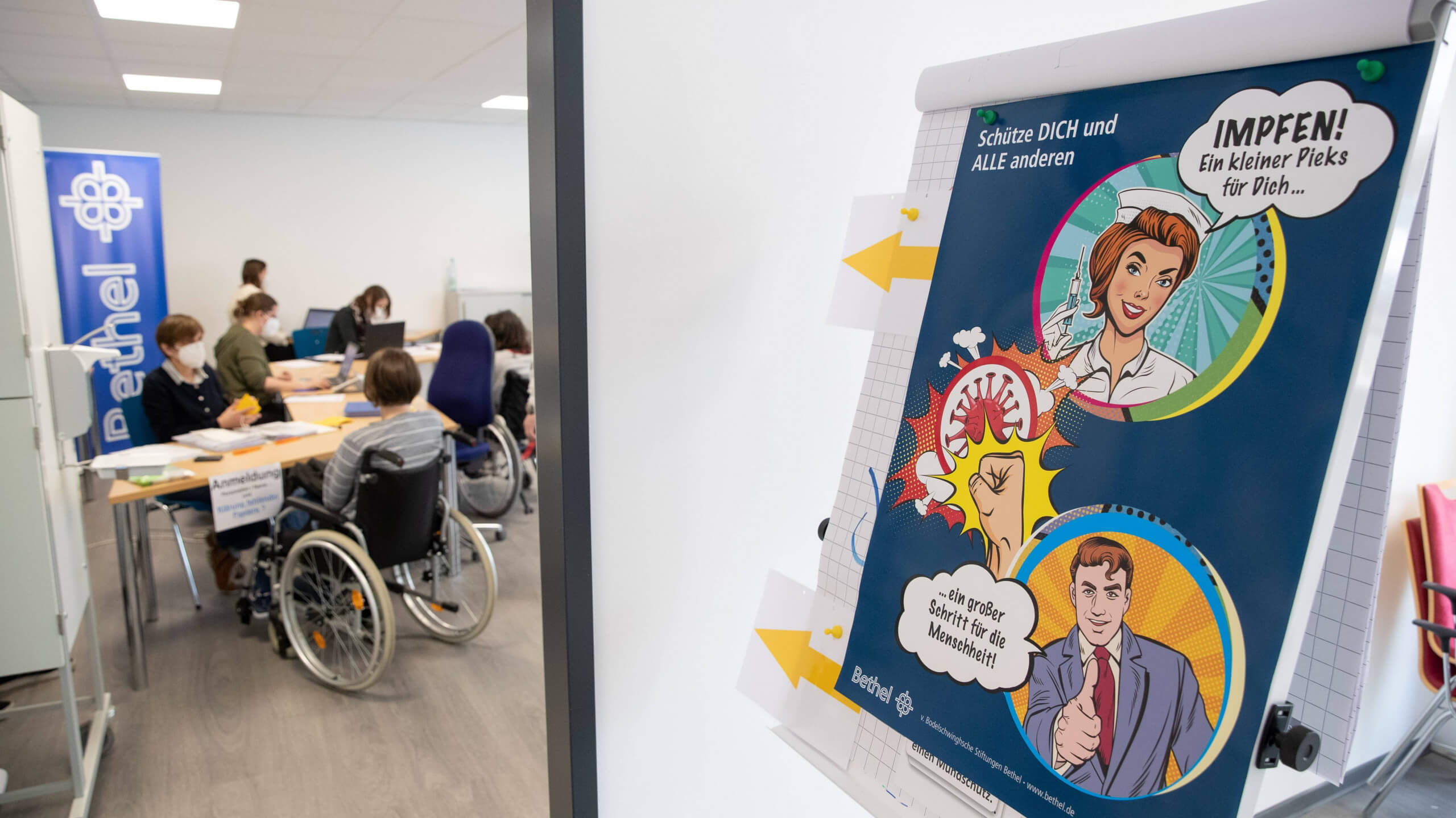 Menschen warten in einem impfzentrum auf eine Corona-Impfung, vor der Tür steht ein impffreundlicher Comic